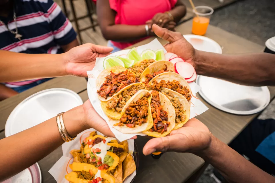 Close-up of multiple hands holding up plate of street-style tacos arranged in circular shape.